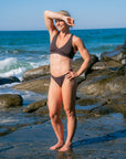 JAYD Swimwear athlete Dom Stitt wearing the cacao Vienna bikini top on a rocky beach with the ocean and blue sky in the background.