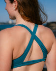 A woman shown from the back wearing the supportive JAYD Swimwear Vienna bikini top in cove featuring crisscrossing straps, set against an outdoor background of blue sky and water.