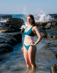 A woman wearing the Vienna bikini top in cove green stands in shallow ocean water on a rocky shoreline, with waves crashing behind her under a clear blue sky.
