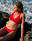 JAYD model wearing a bright red Ruby Vienna bikini top as she sits on dark rocks by the ocean, with waves visible in the background.