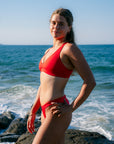 JAYD Swimwear model Jemima wearing the Ruby red Vienna bikini top as she stands on dark rocks by the ocean, with waves and a clear blue sky in the background.