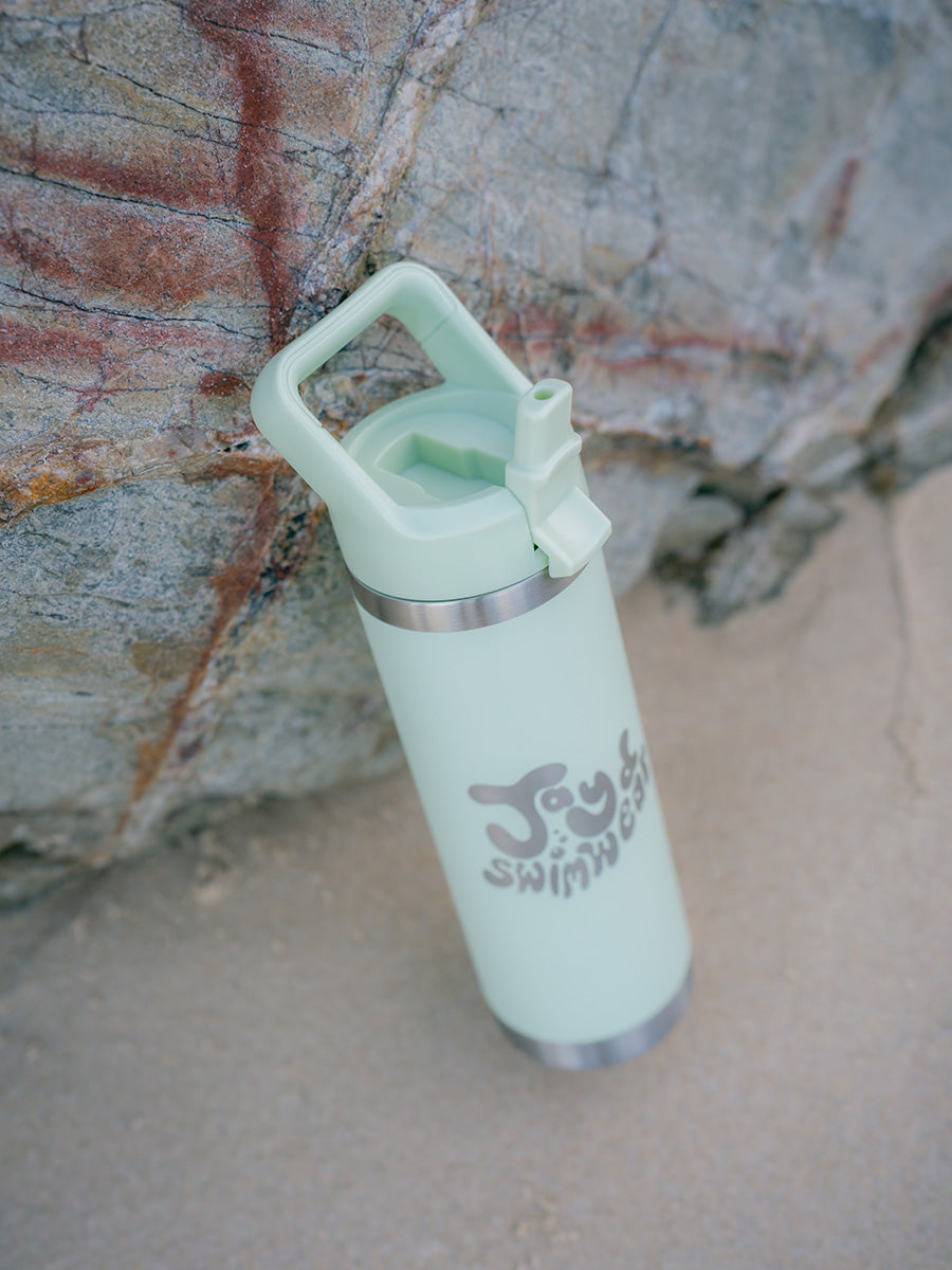 Green water bottle with JAYD Swimwear logo in bubble writing leaning against a rock at the beach.