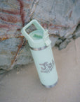 Green water bottle with JAYD Swimwear logo in bubble writing leaning against a rock at the beach.