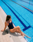 JAYD model in a the Zambia Acai one-piece swimsuit with Tart pink shoulder straps sits on the edge of a swimming pool, looking off-camera. The pool water is bright blue with lane lines, and the deck is light beige tile.