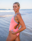 JAYD Swimwear model Chloe wearing the Zambia one-piece swimsuit in vibrant Vibe pink with breeze blue contrasting shoulder straps, on a beach with the ocean waves in the background.