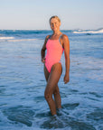 JAYD model Chloe wearing the vibrant Vibe Zambia one piece swimsuit as she stands in the shallow ocean water at a beach, with waves and a pale blue sky in the background.