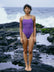 JAYD Swimwear model Sophie standing on a rocky shoreline with white water breaking behind her, wearing the Zambia one piece swimsuit in Murasaki purple with Olive green shoulder straps.