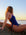 JAYD model sitting on rocks by the ocean at sunset, wearing a Zambia one-piece swimsuit. The swimsuit is navy blue with light purple trim and a small orange accent near the leg opening.