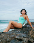 Open water swimmer Madi Armstrong reclining on a rock in front of the ocean wearing the JAYD Swimwear Zambia one piece swimsuit in seamist blue with matcha green straps.
