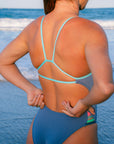 A woman viewed from behind on a beach, wearing the JAYD Swimwear Zambia one piece in storm with light seamist back straps and a multi-panel hip cut-out feature. The ocean waves are visible in the background.