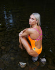 JAYD Model Keira wearing the Zambia one piece in Neon orange, sitting in a shallow stream showcasing the orchid back straps and vibrant hip cut-outs.