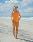 Triathlete Charlotte standing on a beach wearing the JAYD Swimwear Zimmy one piece in Flare orange.