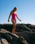  JAYD Swimwear model wearing the bright pink sorbet Zimmy long torso one-piece swimsuit stands barefoot on dark rocky terrain under a clear blue sky.