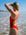 JAYD Swimwear model wearing the bright red Zimmy long torso one-piece swimsuit stands on a sandy beach with ocean waves and a blue sky in the background, photographed from behind.