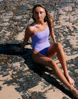 Woman in a Lennox one piece in the colour lilac haze sitting on a rocky beach