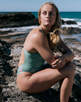 Woman in a matcha green one piece sitting on rocks by the ocean. One piece is made for training