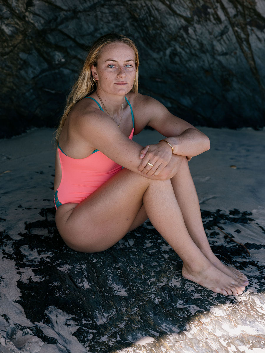 Woman in a JAYD Lennox one piece sitting on a rocky beach. She is wearing the colour vibe with atlantic hip cutouts and strap detailing 