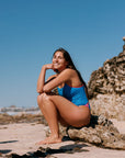 Woman in a cancun blue fixed back one piece sitting on rocks by the water with a clear blue sky.The Zambia one piece has vibe coloured cutouts and straps.