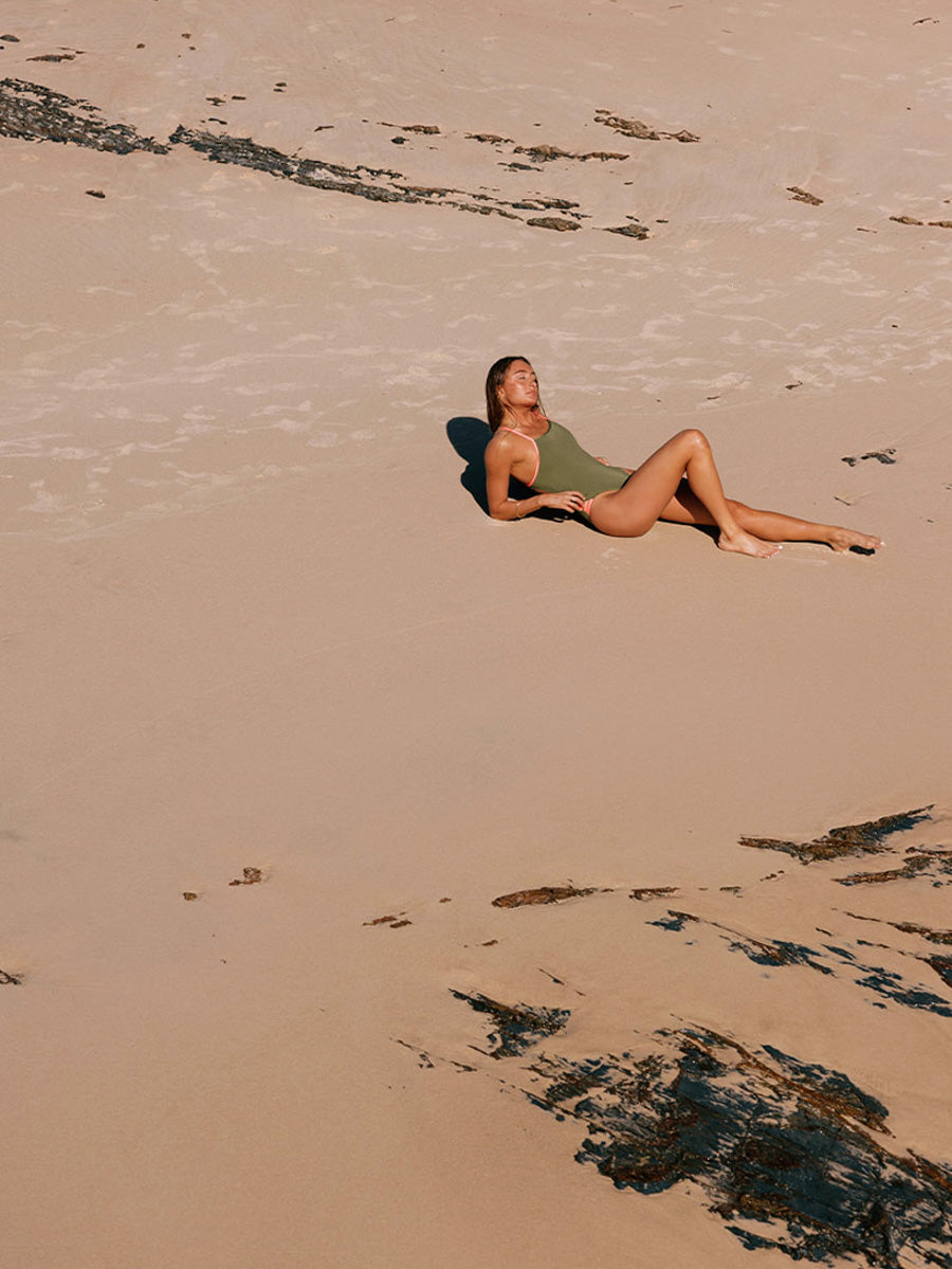 Woman lying on a sandy beach with seaweed scattered around. She is wearing a JAYD swimsuit in the colour Olive