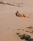 Woman lying on a sandy beach with seaweed scattered around. She is wearing a JAYD swimsuit in the colour Olive
