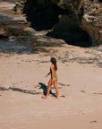 Woman walking on a sandy beach with rocky outcrops. She is wearing a fixed back JAYD one piece built for performance